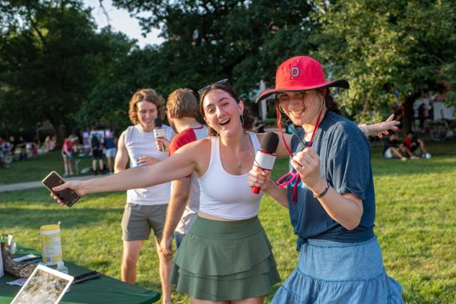 Two female students pose for the camera while two male students talk in the background on campus.